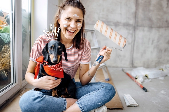 Lachende Person mit Farbspritzern und Hund hält einen Farbroller.